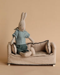 A Maileg Rabbit, Size 2 - Classic Shirt & Shorts, crafted from recycled polyester and dressed in a green shirt and pants, sits on a small beige couch with two back cushions and armrests. The backdrop is a plain beige color, complementing the minimalist and cozy scene reminiscent of a Maileg dollhouse.