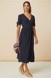 Woman wearing a navy midi dress with short tie sleeves, button front detail, and a front slit, styled with gold wedge espadrilles and a multicoloured handbag.
